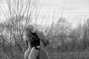 Açık havada mutlu, çarpıcı sarışın kızın moda portresi. Güzel gülümseme. Uzun açık renk saçlı. Şık bir ceket giyiyor. Neşeli ve neşeli bir kadın. Doğal bir arazide, kuru bir sazlığın ve bir gölün yanında yürümek. siyah ve beyaz