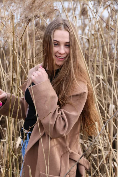 カメラの外でポーズをとっている若い女性 ブラッシュの背景 屋外だ 閉めろ 乾燥した葦と青い湖の近くの晴れた日に 長い髪の美しいブロンド茶色のコートで — ストック写真