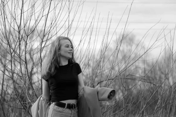 幸せな見事なブロンドの女の子の屋外ライフスタイルのファッションの肖像画 いい笑顔だ 長いライトヘア スタイリッシュなコートを着て 楽しさと陽気な女性 乾燥した葦と湖の近くの自然の風景の上を歩く 黒と白 — ストック写真