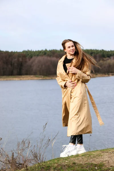 Outdoors lifestyle fashion portrait of happy stunning blonde girl. Beautiful smile. Long light hair. Wearing stylish coat. Joyful and cheerful woman. walk on a natural landscape, near a dry reed and a lake on a sunny day.