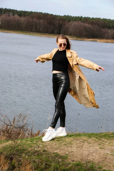 Outdoors lifestyle fashion portrait of happy stunning blonde girl. Beautiful smile. Long light hair. Wearing stylish coat. Joyful and cheerful woman. walk on a natural landscape, near a dry reed and a lake on a sunny day.