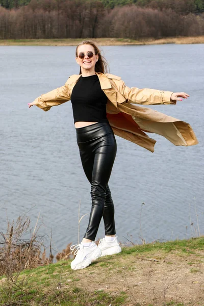 Outdoors lifestyle fashion portrait of happy stunning blonde girl. Beautiful smile. Long light hair. Wearing stylish coat. Joyful and cheerful woman. walk on a natural landscape, near a dry reed and a lake on a sunny day.