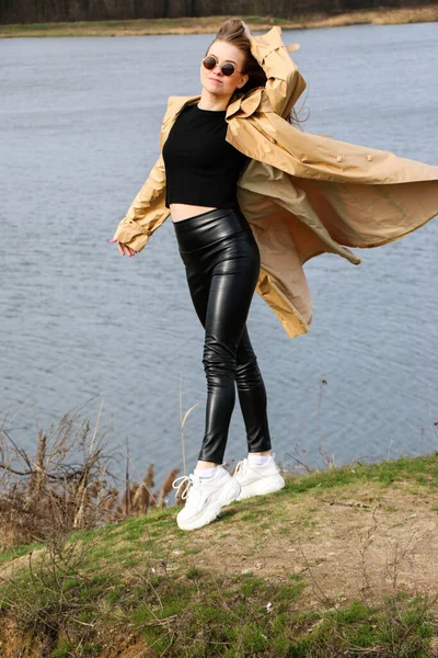 Outdoors lifestyle fashion portrait of happy stunning blonde girl. Beautiful smile. Long light hair. Wearing stylish coat. Joyful and cheerful woman. walk on a natural landscape, near a dry reed and a lake on a sunny day.