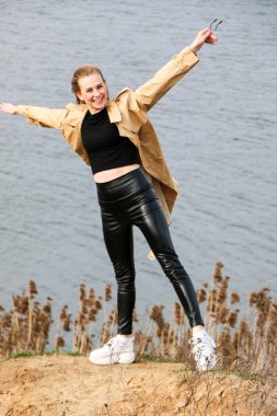 Açık havada mutlu, çarpıcı sarışın kızın moda portresi. Güzel gülümseme. Uzun açık renk saçlı. Şık bir ceket giyiyor. Neşeli ve neşeli bir kadın. Doğal bir arazide yürümek, kuru bir sazlığın yanında ve güneşli bir günde bir göl..