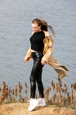 Açık havada mutlu, çarpıcı sarışın kızın moda portresi. Güzel gülümseme. Uzun açık renk saçlı. Şık bir ceket giyiyor. Neşeli ve neşeli bir kadın. Doğal bir arazide yürümek, kuru bir sazlığın yanında ve güneşli bir günde bir göl..