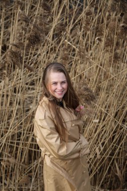 Açık havada mutlu, çarpıcı sarışın kızın moda portresi. Güzel gülümseme. Uzun açık renk saçlı. Şık bir ceket giyiyor. Neşeli ve neşeli bir kadın. Doğal bir arazide, kuru bir sazlığa ve göle yakın yürümek.