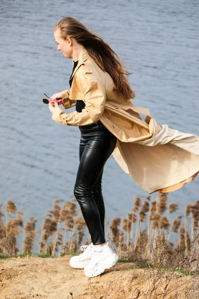 Outdoors lifestyle fashion portrait of happy stunning blonde girl. Beautiful smile. Long light hair. Wearing stylish coat. Joyful and cheerful woman. walk on a natural landscape, near a dry reed and a lake on a sunny day.