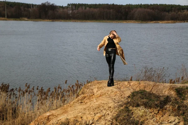 Outdoors lifestyle fashion portrait of happy stunning blonde girl. Beautiful smile. Long light hair. Wearing stylish coat. Joyful and cheerful woman. walk on a natural landscape, near a dry reed and a lake on a sunny day.