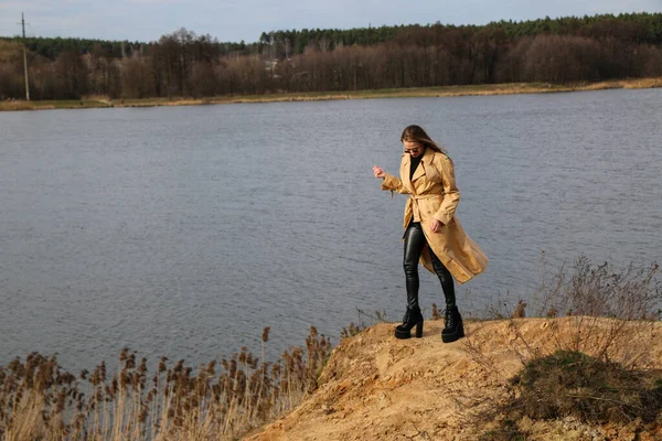 Outdoors lifestyle fashion portrait of happy stunning blonde girl. Beautiful smile. Long light hair. Wearing stylish coat. Joyful and cheerful woman. walk on a natural landscape, near a dry reed and a lake on a sunny day.