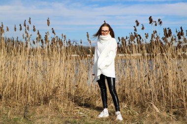 Dışarıdaki kameraya poz veren güzel bir genç kadın. Hızlı bir geçmiş. Açık havada. Kapatın. Kuru sazlığın ve mavi gölün yakınında güneşli bir günde. Kahverengi ceketli, uzun saçlı, güzel bir sarışın.