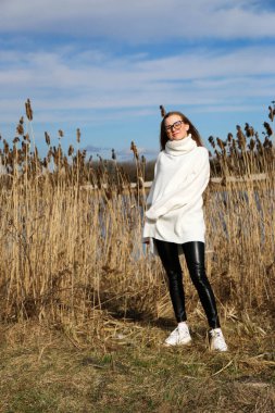 Dışarıdaki kameraya poz veren güzel bir genç kadın. Hızlı bir geçmiş. Açık havada. Kapatın. Kuru sazlığın ve mavi gölün yakınında güneşli bir günde. Kahverengi ceketli, uzun saçlı, güzel bir sarışın.