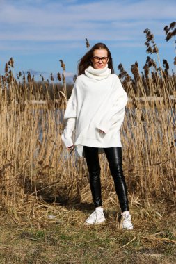 Dışarıdaki kameraya poz veren güzel bir genç kadın. Hızlı bir geçmiş. Açık havada. Kapatın. Kuru sazlığın ve mavi gölün yakınında güneşli bir günde. Kahverengi ceketli, uzun saçlı, güzel bir sarışın.