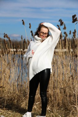 Dışarıdaki kameraya poz veren güzel bir genç kadın. Hızlı bir geçmiş. Açık havada. Kapatın. Kuru sazlığın ve mavi gölün yakınında güneşli bir günde. Kahverengi ceketli, uzun saçlı, güzel bir sarışın.