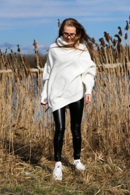 Dışarıdaki kameraya poz veren güzel bir genç kadın. Hızlı bir geçmiş. Açık havada. Kapatın. Kuru sazlığın ve mavi gölün yakınında güneşli bir günde. Kahverengi ceketli, uzun saçlı, güzel bir sarışın.