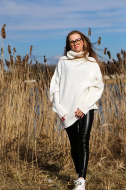 Dışarıdaki kameraya poz veren güzel bir genç kadın. Hızlı bir geçmiş. Açık havada. Kapatın. Kuru sazlığın ve mavi gölün yakınında güneşli bir günde. Kahverengi ceketli, uzun saçlı, güzel bir sarışın.