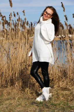 Dışarıdaki kameraya poz veren güzel bir genç kadın. Hızlı bir geçmiş. Açık havada. Kapatın. Kuru sazlığın ve mavi gölün yakınında güneşli bir günde. Kahverengi ceketli, uzun saçlı, güzel bir sarışın.