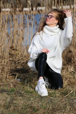 Dışarıdaki kameraya poz veren güzel bir genç kadın. Hızlı bir geçmiş. Açık havada. Kapatın. Kuru sazlığın ve mavi gölün yakınında güneşli bir günde. Kahverengi ceketli, uzun saçlı, güzel bir sarışın.