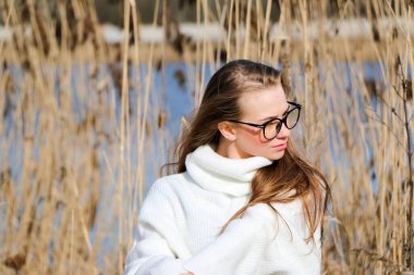 Dışarıdaki kameraya poz veren güzel bir genç kadın. Hızlı bir geçmiş. Açık havada. Kapatın. Kuru sazlığın ve mavi gölün yakınında güneşli bir günde. Kahverengi ceketli, uzun saçlı, güzel bir sarışın.