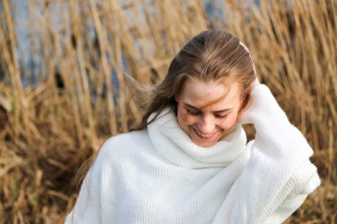 Dışarıdaki kameraya poz veren beyaz kazaklı güzel bir genç kadın. Hızlı bir geçmiş. Açık havada. Kapatın. Kuru sazlığın ve mavi gölün yakınında güneşli bir günde. Kahverengi ceketli, uzun saçlı, güzel bir sarışın.