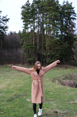 Güzel bir kız, sarışın, uzun saçlı, ilkbaharda ormanın yakınında kahverengi ceketli, dinginlik ve özgürlük içinde yürüyor.