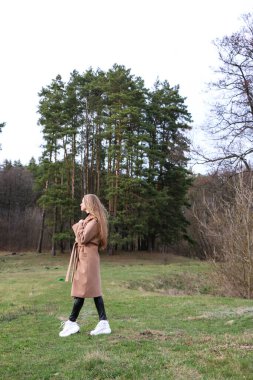 Güzel bir kız, sarışın, uzun saçlı, ilkbaharda ormanın yakınında kahverengi ceketli, dinginlik ve özgürlük içinde yürüyor.