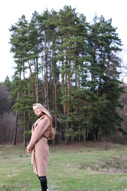 Güzel bir kız, sarışın, uzun saçlı, ilkbaharda ormanın yakınında kahverengi ceketli, dinginlik ve özgürlük içinde yürüyor.