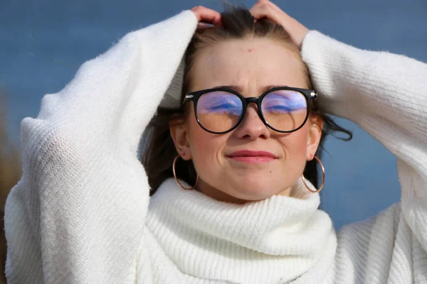 Pretty young woman in a white sweater and glasses posing at camera outdoor. Bulrush background. Outdoor. Close up. On a sunny day near the blue lake. Beautiful blonde with long hair In a brown coat