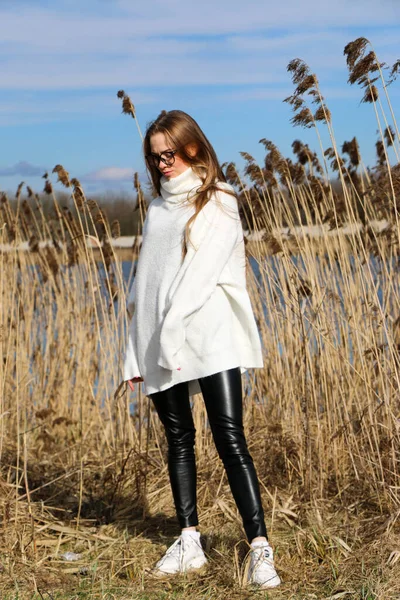 Pretty young woman posing at camera outdoor. Bulrush background. Outdoor. Close up. On a sunny day near the dry reed and blue lake. Beautiful blonde with long hair In a brown coat