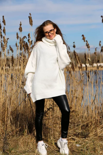 Pretty young woman posing at camera outdoor. Bulrush background. Outdoor. Close up. On a sunny day near the dry reed and blue lake. Beautiful blonde with long hair In a brown coat