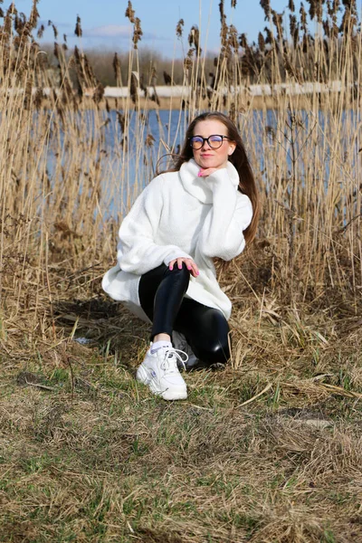 Pretty young woman posing at camera outdoor. Bulrush background. Outdoor. Close up. On a sunny day near the dry reed and blue lake. Beautiful blonde with long hair In a brown coat