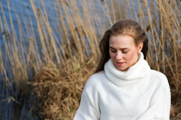 Pretty young woman in a white sweater posing at camera outdoor. Bulrush background. Outdoor. Close up. On a sunny day near the dry reed and blue lake. Beautiful blonde with long hair In a brown coat
