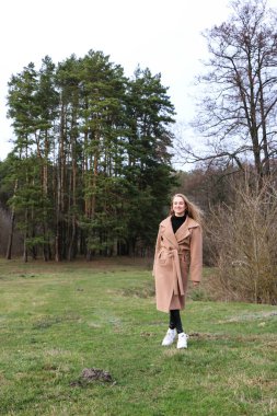 Güzel bir kız, sarışın, uzun saçlı, ilkbaharda ormanın yakınında kahverengi ceketli, dinginlik ve özgürlük içinde yürüyor.