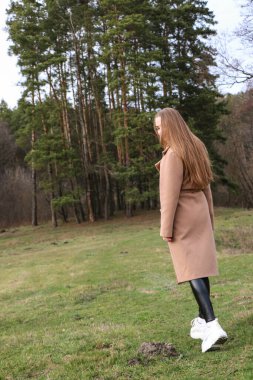 Güzel bir kız, sarışın, uzun saçlı, ilkbaharda ormanın yakınında kahverengi ceketli, dinginlik ve özgürlük içinde yürüyor.