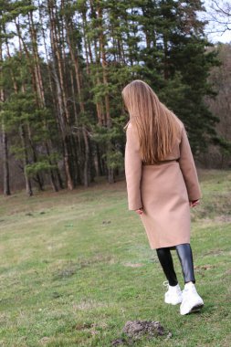 Güzel bir kız, sarışın, uzun saçlı, ilkbaharda ormanın yakınında kahverengi ceketli, dinginlik ve özgürlük içinde yürüyor.