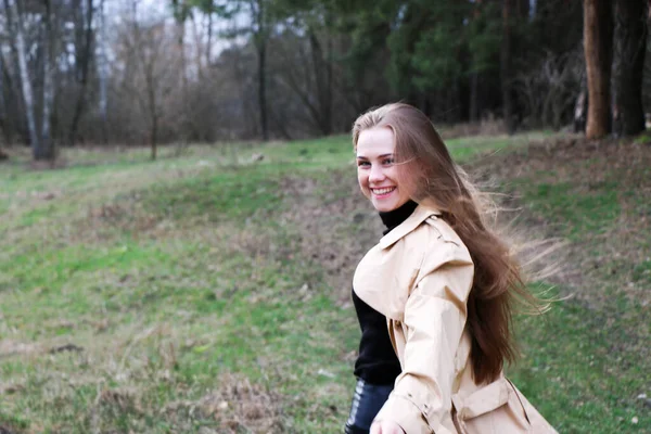 髪の長い金髪の美少女が茶色のコートに身を包んだ森の近くの春を歩く — ストック写真