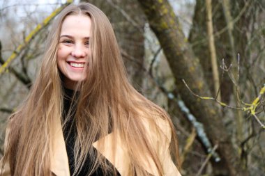 Dışarıdaki kameraya poz veren güzel bir genç kadın. Hızlı bir geçmiş. Açık havada. Kapatın. Kuru sazlığın ve mavi gölün yakınında güneşli bir günde. Kahverengi ceketli, uzun saçlı, güzel bir sarışın.