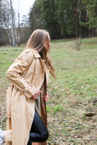 髪の長い金髪の美少女が茶色のコートに身を包んだ森の近くの春を歩く — ストック写真