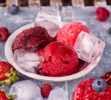 Ev yapımı çilek, kuşüzümü, ahududu, yabanmersinli dondurma kasede şerbet, kepçe, böğürtlen ve buz küpleri mavi ahşap arka planda. Yaz tatlısı, tatlı atıştırmalık. 