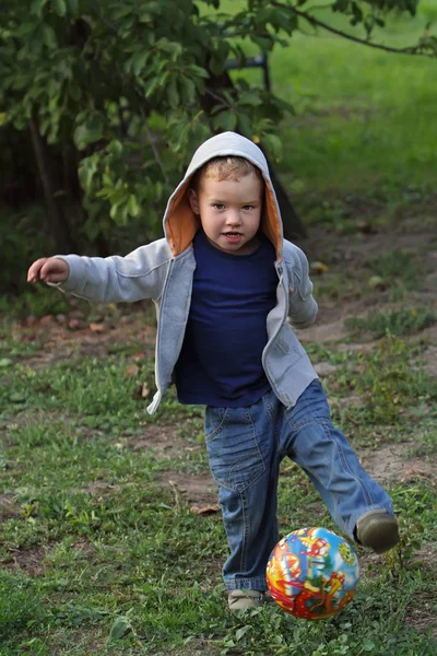 A little boy in jeans clothes plays with a bright ball in football. A funny kid in a hood holds a bright round ball. — Stock Photo, Image