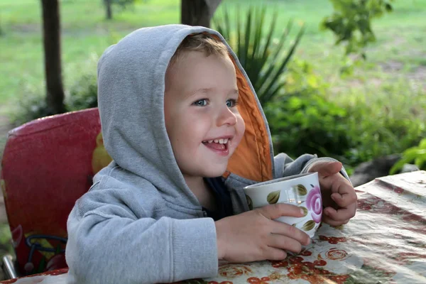 A little boy in a gray hood is drinking from a cup. Emotional ki — Stock Photo, Image