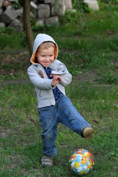 Kot pantolon giysili küçük bir çocuk futbol parlak bir top ile oynuyor. Eğlenceli çocuk sesle gülüyor. — Stok fotoğraf