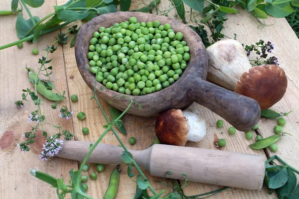 A beautiful still-life in the rustic style consists of their green fresh peas in old wooden utensils, a large white edible mushroom, a pistil and green shoots spread out on a rough wooden table. Green raw vegetables and plants a huge supply of energy