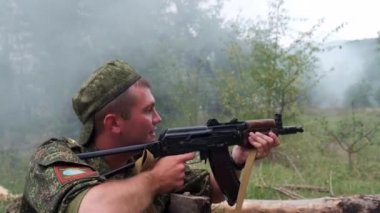 Rusya, Maykop - 2 Ağustos 2019. Bir asker siperde oturuyor, Ak-74 askeri silahlarının düşmanlarına ateş ediyor. Ormanda bir düşman askeri ile savaşmak. Duman ekranı
