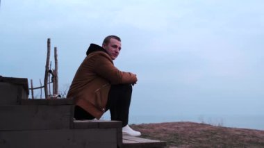 Beyaz takım elbiseli, siyah kot pantolonlu ve beyaz ayakkabılı genç beyaz adam ahşap merdivenlerde oturuyor. Denize bakan yüksek bir dağda, evin yakınında otlar yetişir. Akşam vakti, mavi gökyüzü ile gün batımı.