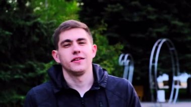 Siyah ceketli beyaz bir adam parkta yürür ve kamerayla sabit bir alanda konuşur. Mutlu bir adamın konuşması ön planda. Dışarıda bir adam gülümsüyor. Yağmurlu hava.