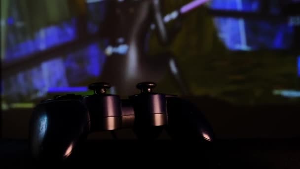 ゲームコンソール上でビデオゲームをプレイするブラックコントローラー 大画面 プロジェクターやテレビの前でゲームパッド カメラはテーブルに沿って移動します パノラマのシーンをクローズアップ表示します 緑への光の変化 — ストック動画