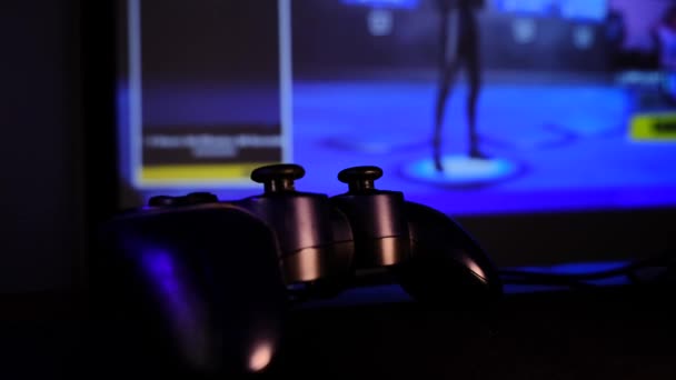 ゲームコンソール上でビデオゲームをプレイするブラックコントローラー 大画面 プロジェクターやテレビの前でゲームパッド カメラはテーブルに沿って移動します パノラマのシーンをクローズアップ表示します 緑への光の変化 — ストック動画