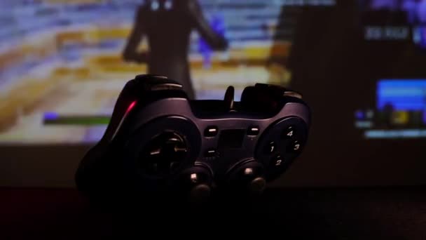 Black controller to play video games on game console. Gamepad in front of a large screen, projector or TV. Camera moves along table. Panoramic scene close-up view. Light changes to blue, red, green. — Stock Video