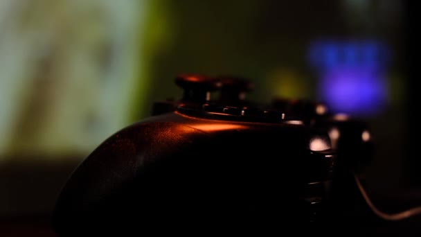 Közelkép kilátás fekete vezérlő az asztalon játszani videojátékok játékkonzol. Gamepad előtt egy nagy képernyő, projektor vagy TV. A lámpa kék, piros, zöld színre változik.. — Stock videók