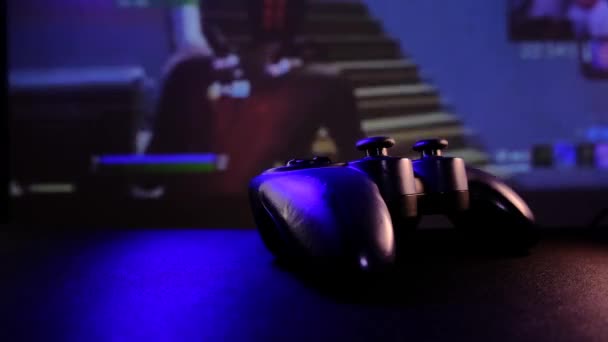テーブルの上のクローズアップビューブラックコントローラーは、ゲームコンソール上でビデオゲームをプレイします。大画面、プロジェクターやテレビの前でゲームパッド。ライトランプは青、赤、緑に変わります. — ストック動画